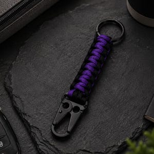 Könnte beinhalten: Ein Schlüsselanhänger mit einem geflochtenen Paracord-Design in abwechselnden Farben Schwarz und Violett. Die Kordel ist an einem schwarzen Metall-Spaltring oben und einem schwarzen Metall-Karabinerhaken unten befestigt, um Schlüssel sicher zu halten.