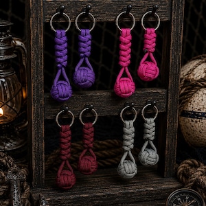 Op de afbeelding: Acht paracord sleutelhangers met apenvuistknopen, gerangschikt in paren van paars, roze, bordeauxrood en grijs. Elke sleutelhanger bestaat uit een gevlochten koordgedeelte bevestigd aan een metalen ring, hangend aan een houten displayrek.