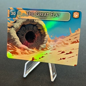 Puede incluir: Una carta de juego de cartas azul y naranja con el texto "The Great Flat" y "Arrakis" impreso en ella. La carta presenta un gran agujero circular oscuro en el suelo con formaciones de arena y roca que lo rodean. La carta tiene el número "30" impreso en la esquina superior izquierda.