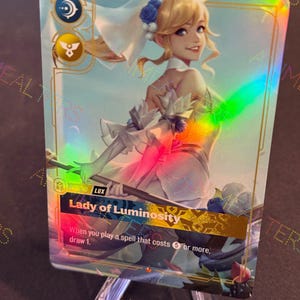 May include: A holographic trading card featuring a blonde woman in a white and gold dress, with the text "Lady of Luminosity." The card has a rainbow sheen and is displayed on a clear stand. The card's text reads, "When you play a spell that costs 6 or more, draw 1."
