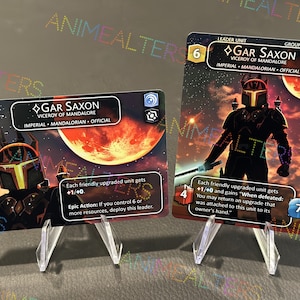Puede incluir: Dos cartas coleccionables con Gar Saxon, Virrey de Mandalore, con un fondo temático espacial. Las cartas muestran arte de personajes, estadísticas y texto del juego. Una carta es una Unidad de Líder con un 6, y la otra tiene un 4 y un 7.