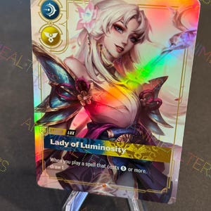Può includere: Una carta collezionabile olografica raffigurante una donna con lunghi capelli bianchi e un copricapo floreale. La carta ha una lucentezza arcobaleno e il testo "Lady of Luminosity". La carta è esposta su un supporto di plastica trasparente.