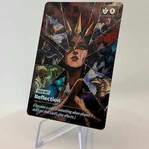 Puede incluir: Una carta coleccionable rectangular titulada Reflection, que muestra una figura central con corona rodeada de ilustraciones fragmentadas de varios personajes. La carta incluye el texto Token Unit y una descripción de su habilidad.