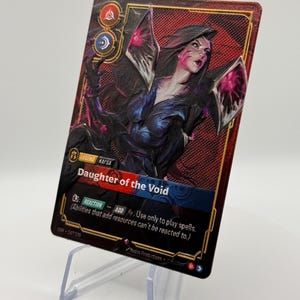 May include: A trading card featuring a female character with dark hair and pink accents, titled "Daughter of the Void." The card has a red and black background with text that reads "REACTION - ADD." The card is displayed on a clear plastic stand.
