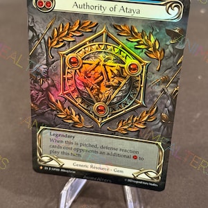 Puede incluir: Una carta coleccionable titulada "Authority of Ataya" con un emblema dorado y rojo. La carta presenta una ilustración detallada con el texto "Legendary" y "Generic Resource - Gem". La carta se muestra en un soporte transparente.