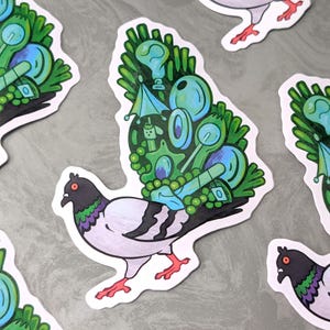 May include: A collection of stickers featuring a stylized pigeon design. The pigeon has a black head, white body, and red feet. The wings are decorated with green and blue illustrations of light bulbs, spoons, and other objects. The stickers are on a gray surface.