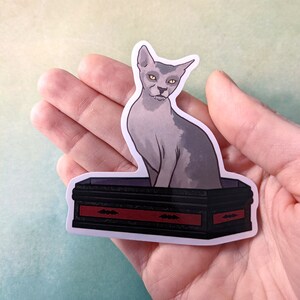 May include: A sticker featuring a grey cat with yellow eyes sitting on a black coffin with a red trim and bat designs. The coffin is decorated with a red trim and bat designs.