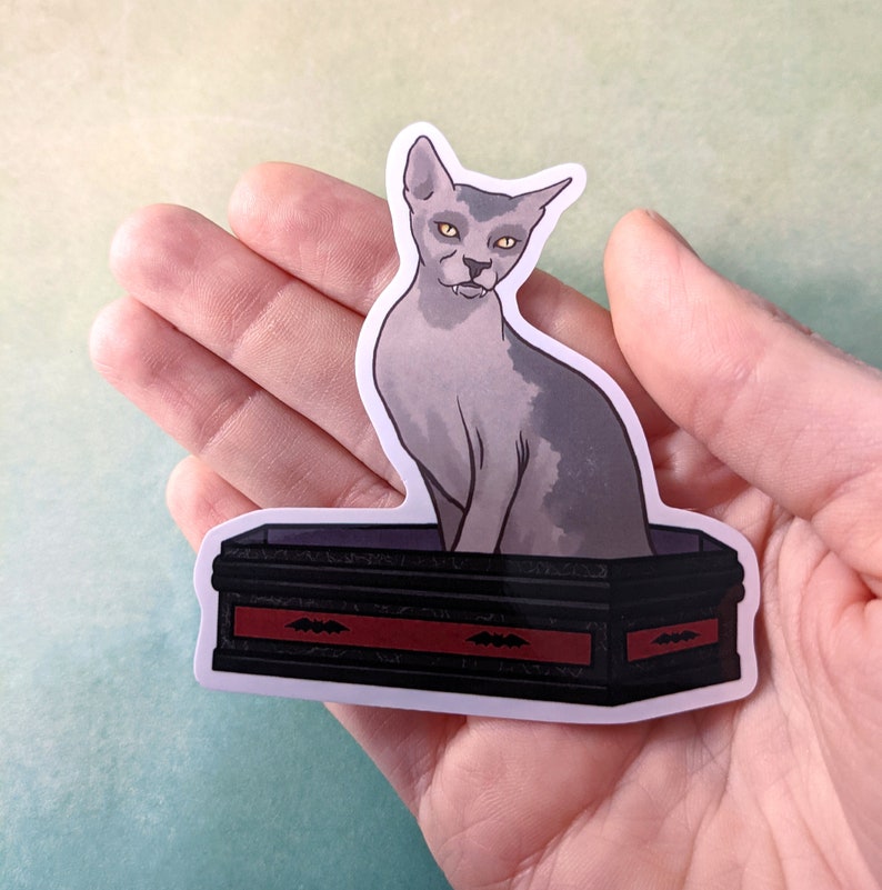 May include: A sticker featuring a grey cat with yellow eyes sitting on a black coffin with a red trim and bat designs. The coffin is decorated with a red trim and bat designs.