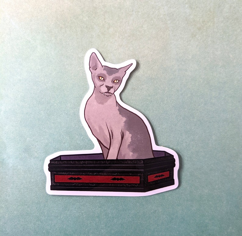 May include: A sticker featuring a grey hairless cat sitting in a black coffin with red trim and bat designs. The cat has yellow eyes and a serious expression.