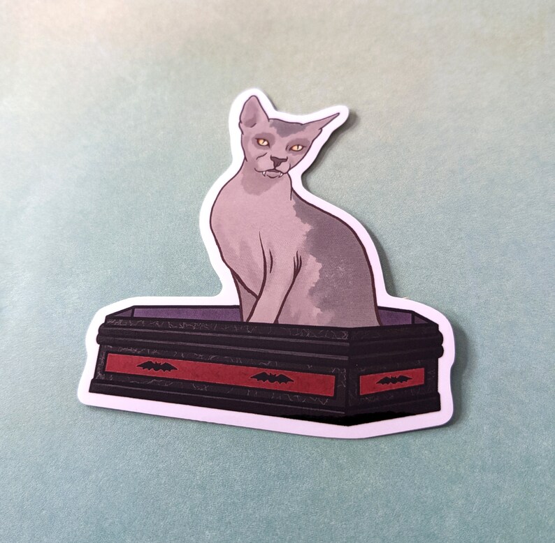 May include: A grey and white cat with yellow eyes sits in a black coffin with red trim and bat designs. The coffin is on a light blue background.