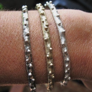 May include: Three silver bracelets with textured surfaces and small round beads. The bracelets are stacked on a person's wrist.