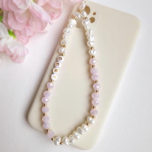 May include: A white phone case with a pink and white beaded phone strap. The strap has gold beads and the letters "GERY" spelled out in gold letters.