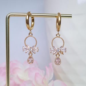 May include: Gold hoop earrings with a dangling charm featuring a circle with a bow made of clear cubic zirconia stones and a teardrop-shaped stone.