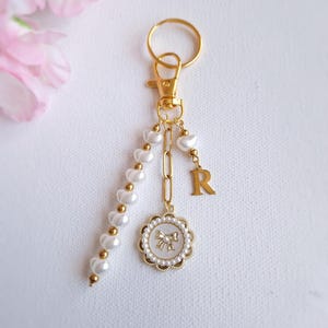 Może przedstawiać: Złoty brelok do kluczy z zawieszką z pereł i sercem, małą zawieszką w kształcie kokardki i zawieszką z literą 'R'.