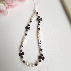 Può includere: Un ciondolo per telefono in perle bianche e nere con un accento dorato e fiocchi neri. Il ciondolo ha il nome "AVA" scritto in lettere bianche.