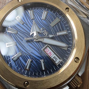 Puede incluir: Reloj de pulsera de acero inoxidable y dorado con esfera azul estampada. El reloj tiene una pantalla de fecha que muestra "DOM 5" y agujas e indicadores luminosos. El reloj tiene una correa de acero inoxidable.