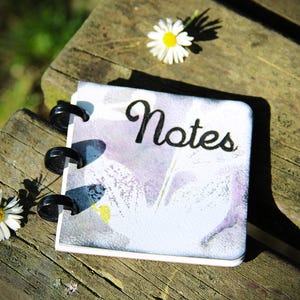 Może przedstawiać: Mały notatnik w spiralnej oprawie z napisem "Notes" czarnym pismem na okładce. Okładka ma akwarelowy wzór kwiatowy w odcieniach fioletu i bieli. Dwa stokrotki umieszczono obok notatnika na drewnianej powierzchni.