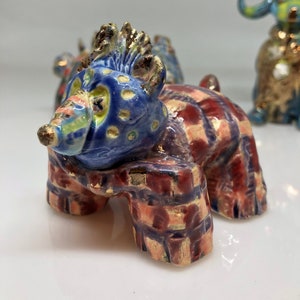 May include: A ceramic figurine of a blue and red bear with gold accents. The bear has a spiky mohawk and is wearing a plaid scarf.
