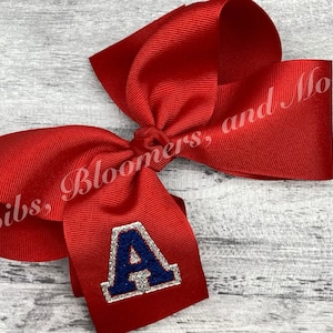 May include: A red hair bow with a silver and blue glitter letter A in the center. The bow is on a white wooden background.