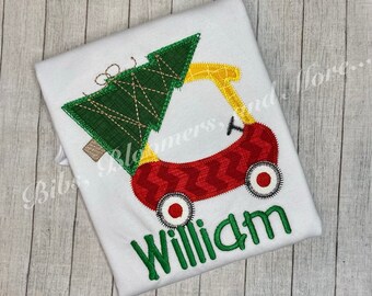 May include: A white t-shirt with a red wagon carrying a green Christmas tree. The wagon has yellow wheels and a red and white chevron pattern. The name "William" is embroidered in green below the wagon.