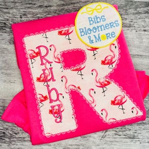 May include: Hot pink shirt with a large, embroidered letter "R" and the name "Ruby" in pink thread. The letter is filled with a flamingo pattern and framed with a white lace trim. A circular patch with the text "Bibs Bloomers & More" is in the upper right.