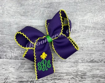 Könnte beinhalten: Eine violette Haarschleife aus Ripsband mit leuchtend gelben Ziernähten an den Kanten. Die Mitte ist mit einem grünen Band gerafft. Ein gelbes Fleur-de-Lis-Emblem ist über den grünen Monogrammbuchstaben gRE auf einem Bandende eingestickt.