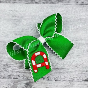 May include: A green and white bow with a red and white candy cane letter "A" embroidered on it. The bow has a white lace trim.
