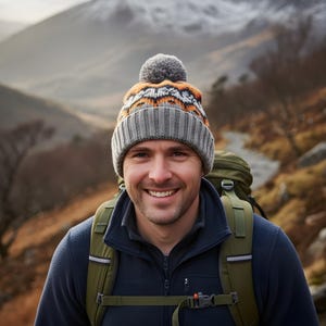 May include: A grey, orange, and white knitted beanie hat with a pom-pom. The person is wearing a navy blue fleece jacket and a green backpack. The background shows a mountain range.