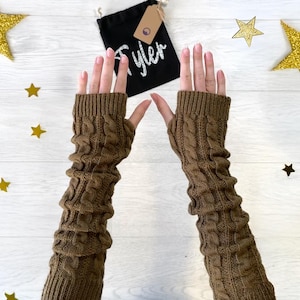 Könnte beinhalten: Ein Paar braune gestrickte Armwärmer mit einem Zopfmuster. Die Armwärmer sind lang und reichen bis zum Ellbogen.