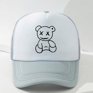 May include: A white and light blue trucker hat with a black and white cartoon bear with an 'X' for eyes. The bear is sitting down.