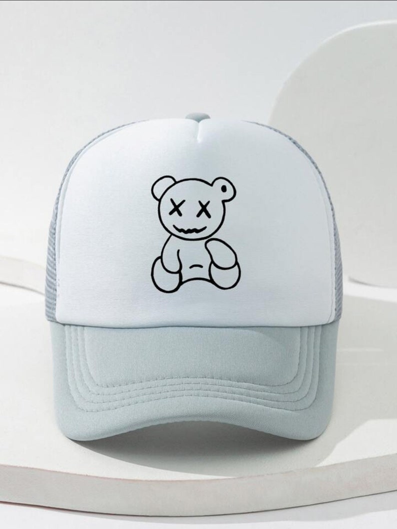May include: A white and light blue trucker hat with a black and white cartoon bear with an 'X' for eyes. The bear is sitting down.