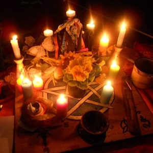 Puede incluir: Una escena con poca luz que presenta una mesa de madera adornada con velas de varios colores y tama&ntilde;os. Un pentagrama est&aacute; pintado sobre la mesa, rodeado de flores, un cuenco peque&ntilde;o y otros objetos rituales. Las velas est&aacute;n encendidas, creando un brillo c&aacute;lido.