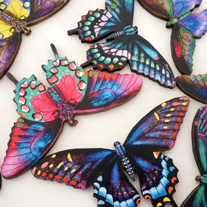May include: A collection of colorful butterfly decorations with intricate wing patterns. The butterflies feature a variety of colors, including blue, green, red, and yellow, with detailed designs. The wooden butterflies are arranged on a white surface.