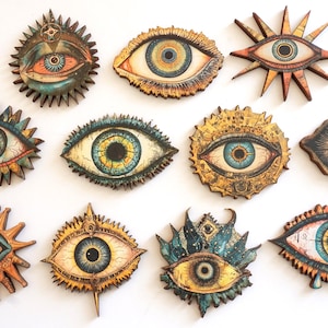 Op de afbeelding: Twaalf decoratieve houten oogwanddecoraties met ingewikkelde details en levendige kleuren. Elk oog is omgeven door een uniek ontwerp, waaronder sterren, bloemen en geometrische patronen. De ogen zijn geschilderd in tinten blauw, groen, geel en oranje.