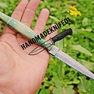 May include: A handmade knife with a green handle and a patterned blade is held in a hand. The knife has a black handle and the text "HANDMADEKNIFE01" is visible.
