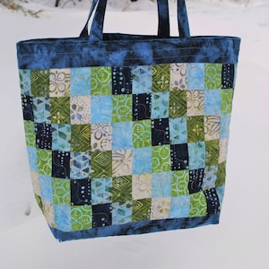 May include: A blue patchwork tote bag with green, white, and blue fabric squares. The bag has two blue straps and is hanging from a branch in a snowy winter scene.