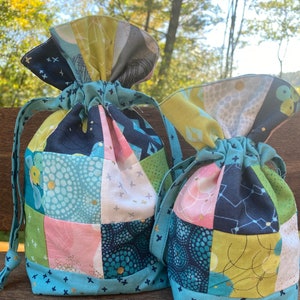 May include: Two fabric drawstring bags with patchwork designs in shades of blue, green, pink, and yellow. The bags are made of different patterned fabrics sewn together in a square grid pattern. The bags have a blue and white trim with gold accents.