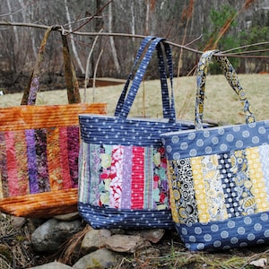 May include: Three tote bags made from colorful patchwork fabric. The bags have blue, yellow, orange, purple, pink, and green fabric strips. The bags have long handles and are sitting on a bed of brown leaves and rocks.