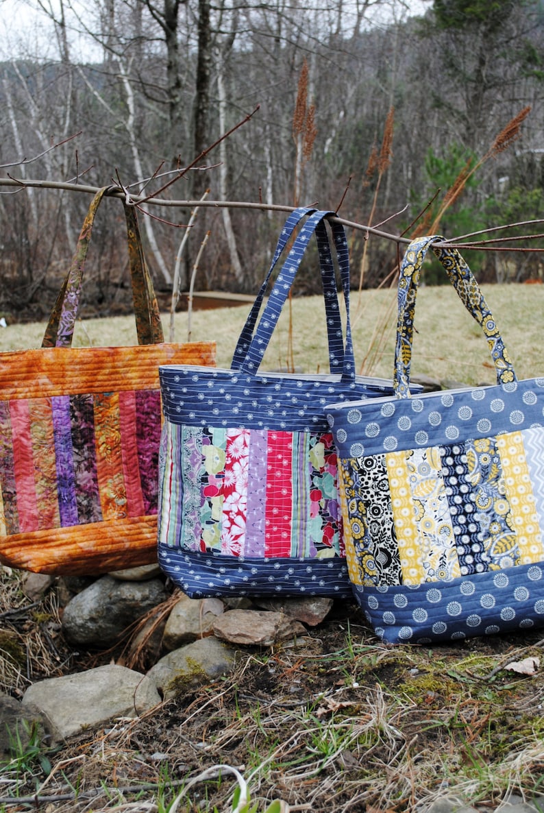 May include: Three tote bags made from colorful patchwork fabric. The bags have blue, yellow, orange, purple, pink, and green fabric strips. The bags have long handles and are sitting on a bed of brown leaves and rocks.