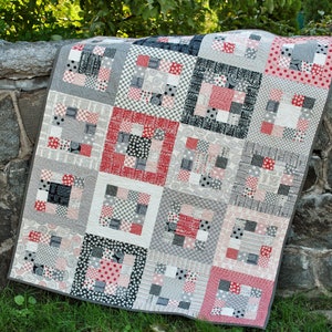 May include: A patchwork quilt in a red, white and grey colour scheme. The quilt is made up of squares of different fabrics, each with a unique pattern. The quilt is folded and draped over a stone wall.