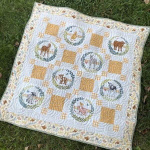 May include: A white and yellow patchwork quilt with a farm animal theme. Each square features a different farm animal surrounded by a wreath of green leaves. The quilt has a white and yellow checkered border.