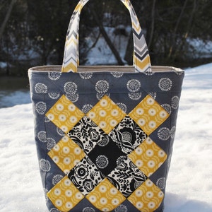 Peut inclure: Un sac fourre-tout en patchwork gris et jaune avec une poignée à motif chevron. Le sac est en tissu et présente un design matelassé.