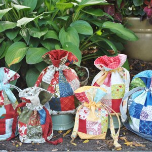 Peut inclure: Six sacs &agrave; cordon en patchwork color&eacute;s, faits de chutes de tissu dans diff&eacute;rents motifs et couleurs, notamment rouge, bleu, vert, jaune et blanc. Les sacs sont dispos&eacute;s sur une surface brune avec des plantes vertes en arri&egrave;re-plan.