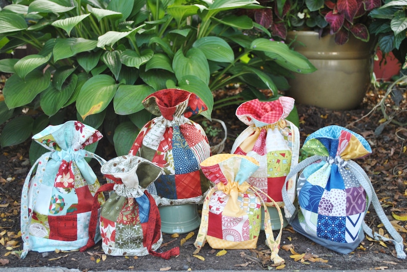 Peut inclure: Six sacs &agrave; cordon en patchwork color&eacute;s, faits de chutes de tissu dans diff&eacute;rents motifs et couleurs, notamment rouge, bleu, vert, jaune et blanc. Les sacs sont dispos&eacute;s sur une surface brune avec des plantes vertes en arri&egrave;re-plan.