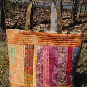 May include: A large, quilted tote bag with a patchwork design. The bag is made of various shades of brown, pink, purple, and blue fabric. The bag has two long straps and a wide base.