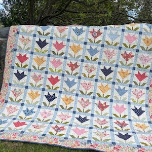 May include: A handmade quilt featuring a repeating tulip pattern in various colors, including pink, blue, yellow, and red. The quilt has a white background with a blue grid pattern and a floral border. The quilt is displayed outdoors on a stone wall.