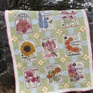 May include: A quilted blanket featuring nine panels with vintage seed packet designs. The panels showcase various flowers like zinnias, delphiniums, sunflowers, cosmos, sweet peas, California poppies, marigolds, and pansies. The quilt has a green and white checkered pattern.