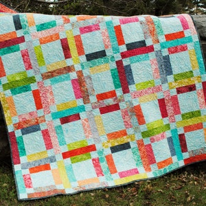 May include: A colourful quilt with a patchwork design featuring various shades of red, orange, yellow, green, blue, and turquoise. The quilt is made of fabric strips sewn together in a grid pattern. The quilt is hanging on a tree branch.