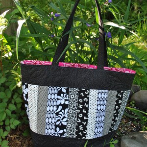 May include: A black and white patchwork tote bag with a pink lining. The bag has a variety of patterns, including polka dots, stripes, and floral designs. The bag is sitting on a bed of rocks and green plants.