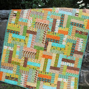 May include: A patchwork quilt with a variety of colorful fabrics, including green, blue, orange, and brown. The quilt is made up of squares and rectangles of different sizes and patterns. The quilt is lying on a bed of green grass.